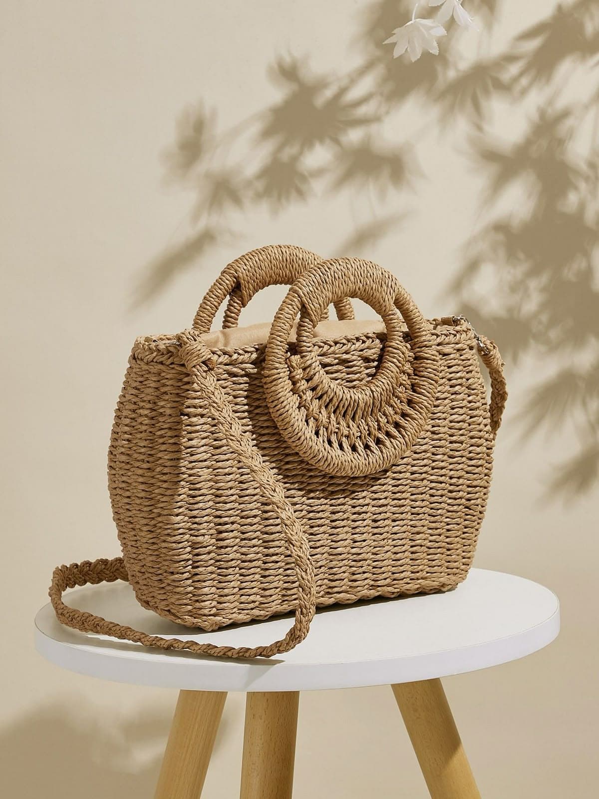 Woven straw handbag with round handles and a braided shoulder strap on a white stool.